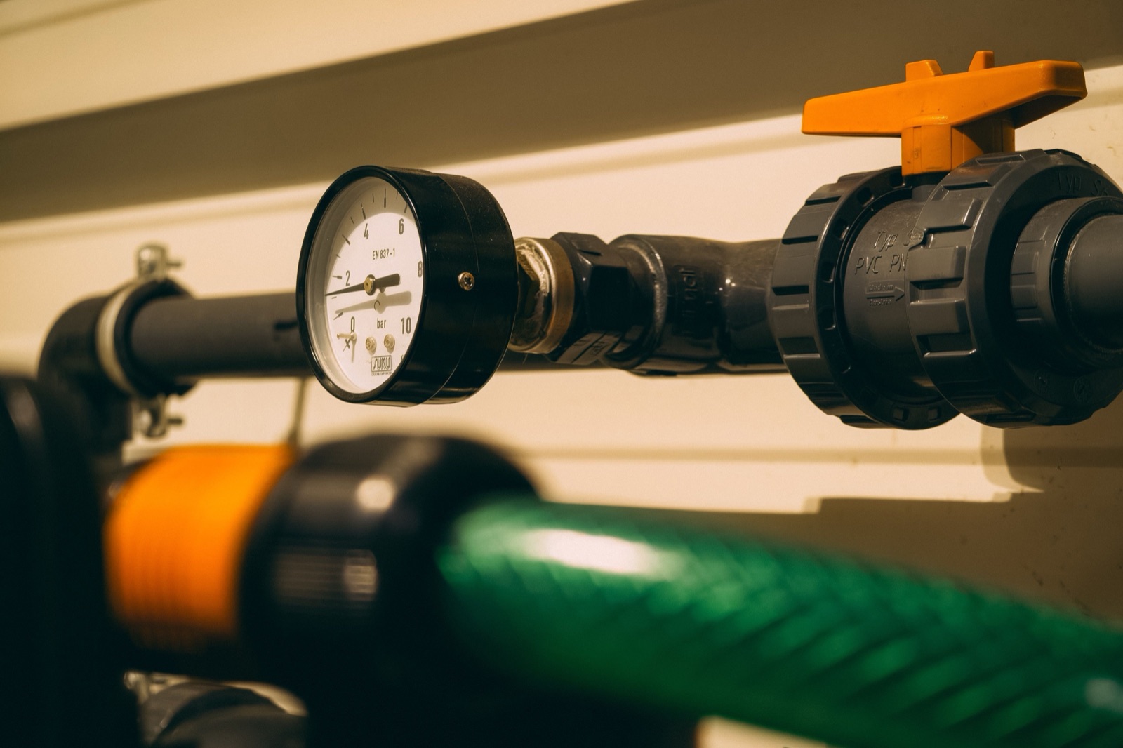 Manometer an einer grauen Kunststoffleitung mit orangefarbenem Kugelhahn und unscharfem grünem Schlauch in einem Technikraum, wo sich Leitungen erwärmen und Legionellen im Kaltwasser begünstigen können