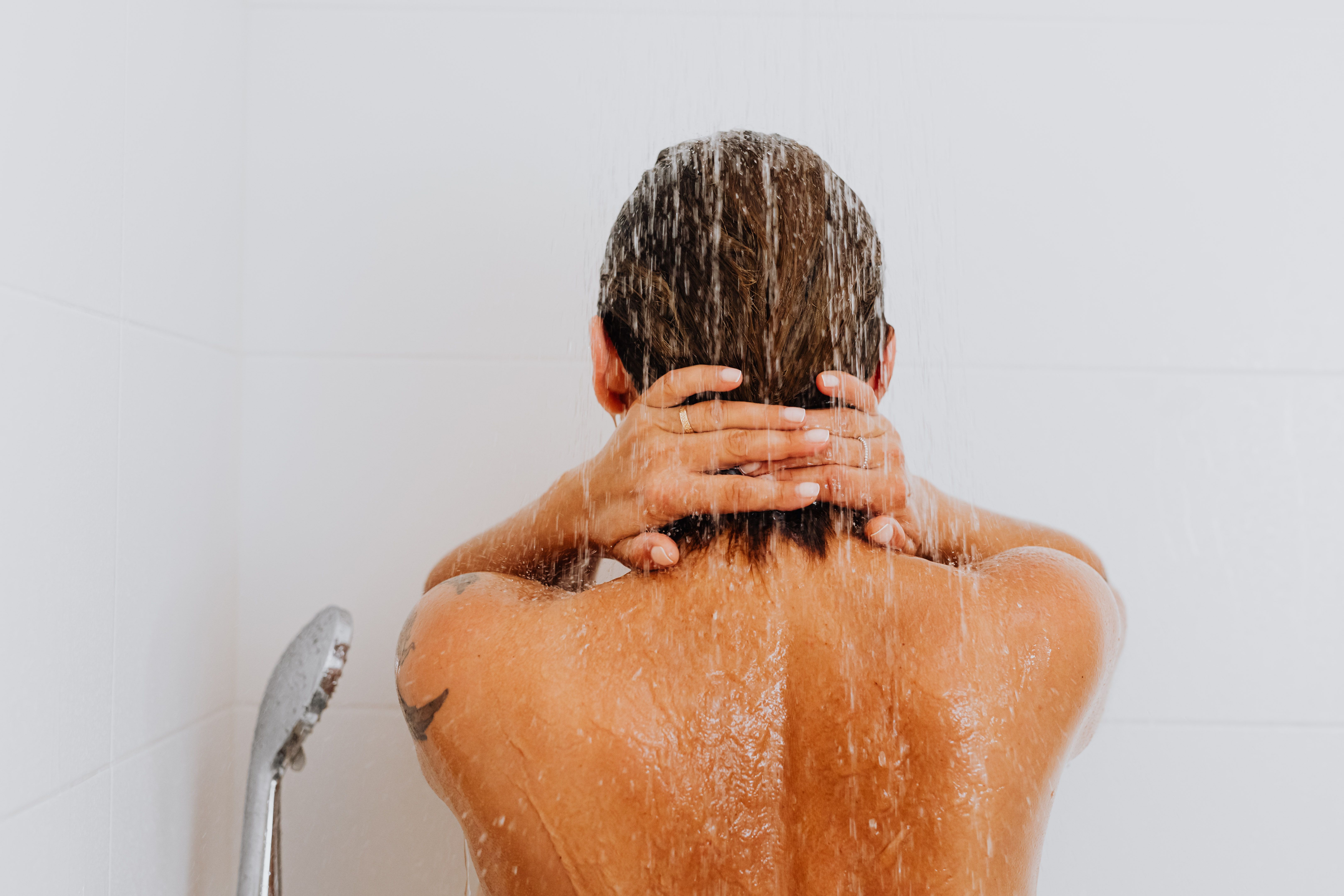Person von hinten unter der laufenden Dusche, das Wasser läuft über Haare und Rücken
